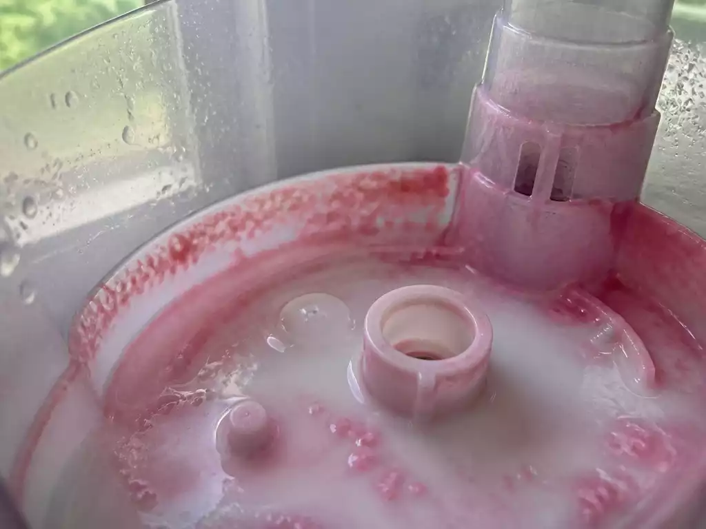 Close-up view of pink residue inside a humidifier tank, showing slimy bacterial growth on the plastic surface, with water droplets, natural light