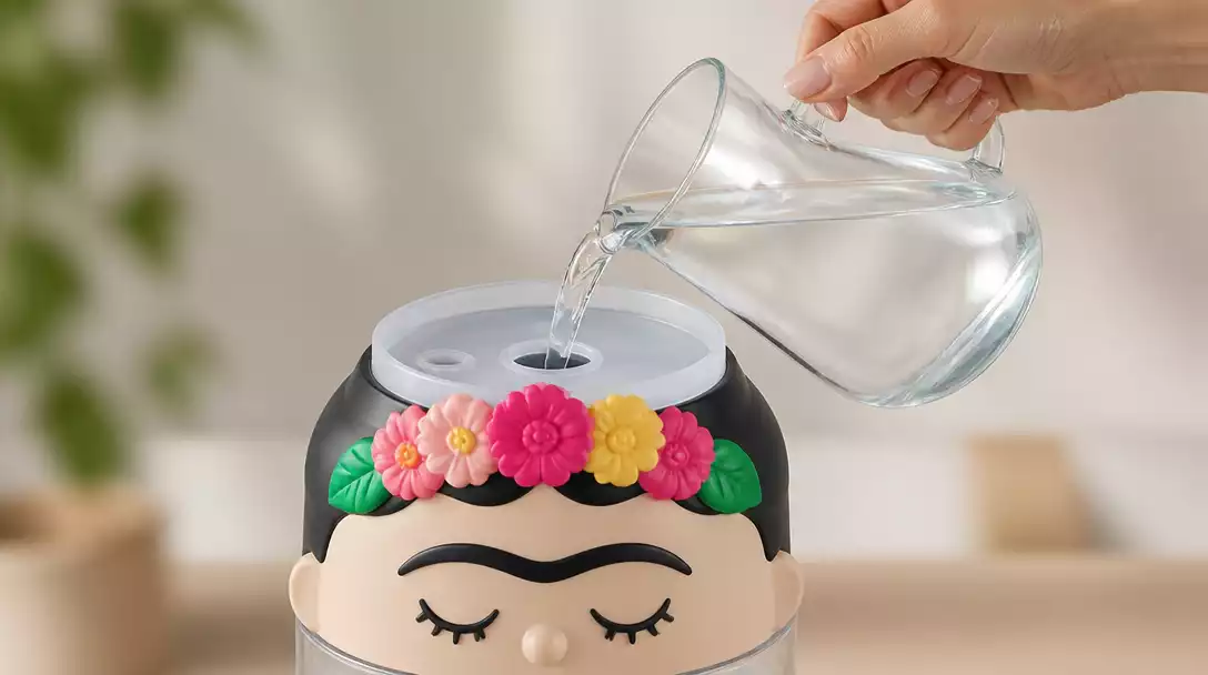 Close-up of a Frida Humidifier being refilled with water, showing the top-fill design and a hand holding a water pitcher