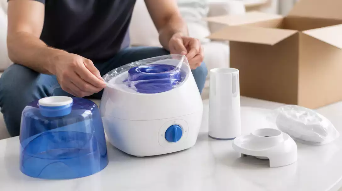 Person unboxing a Vicks humidifier, carefully removing protective plastic and tape, with components like the water tank and mist chimney visible